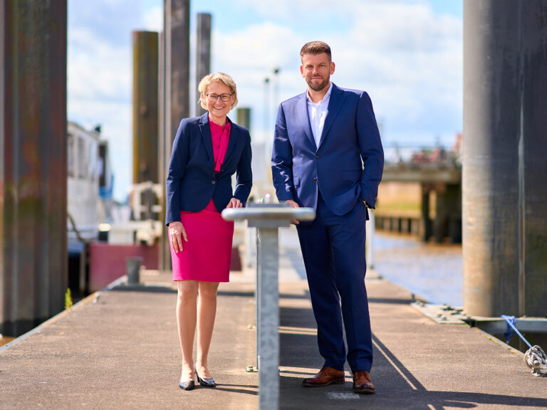Christiane Thürlings in einem blauen Blazer und rosa Kleid, steht links an einem Tisch. Rechts steht Sebastian Ost in einem blauen Anzug und weißem Hemd. Im Hintergrund ist verschwommen der Hafen von Brake zu sehen.